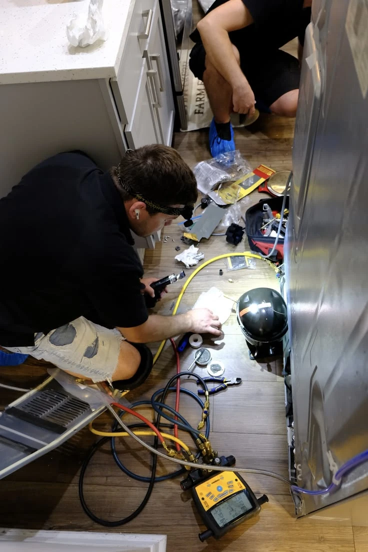 Technicians checking refrigerator components with hoses, gauges, and diagnostic equipment
